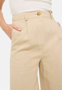 Beige højtaljede bukser med en afslappet pasform, lavet af et tekstureret stof, med frontfald og en enkelt knaplukning.