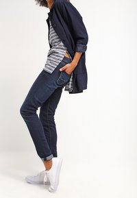 Person som bär mörkblå jeans uppvikta vid ankeln, vita sneakers, randig skjorta och marinblå knäppt jacka med uppvikta ärmar.