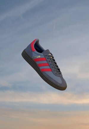 Sapatilha desportiva cinza com acentos vermelhos, três riscas e logotipo dourado. Apresenta uma sola em borracha e parte superior texturizada para aderência e durabilidade.