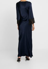 Vestido maxi de satén en color navy con mangas largas que presenta flecos negros en los puños, textura suave y un drapeado relajado. Se muestran unos tacones con abertura en la parte trasera.