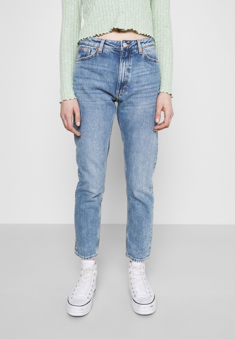 Jeans en denim bleu clair à coupe droite, finition délavée, design classique à cinq poches, associés à des baskets blanches et un haut côtelé vert.
