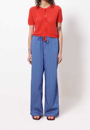 Persona que lleva una camiseta roja de manga corta con textura y lazos delanteros, pantalones anchos azules con rayas verticales rojas y sandalias rojas con los dedos descubiertos.