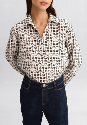 Donna che indossa una camicia bianca con bottoni e stampa floreale nera infilata in un paio di jeans blu scuro, su sfondo semplice.