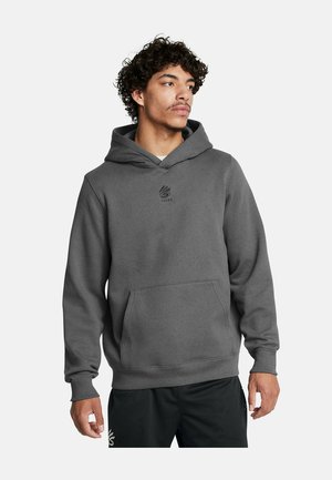 Giovane uomo che indossa una felpa con cappuccio grigia scura e pantaloni corti in rete nera, che guarda di lato su uno sfondo neutro.