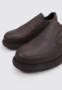 Zapatos de piel marrón tipo mocasín con una textura lisa, puntera redondeada y costuras en contraste. Suela de goma negra gruesa para mayor comodidad.