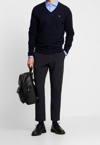 Marinblå v-ringad tröja, ljusblå skjorta med krage, mörka byxor, svart läderryggsäck med teal-accenter och svarta snörskor.