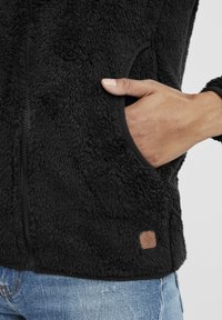Schwarze Fleecejacke mit einer weichen, strukturierten Oberfläche. Verfügt über einen Reißverschluss, seitliche Taschen und ein kleines braunes Logoetikett am Saum.