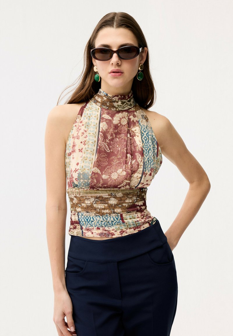 Femme portant un haut sans manches à col haut et motif, des lunettes de soleil noires, des boucles d'oreilles pendantes vertes et un pantalon bleu marine, posant avec une main sur la hanche.