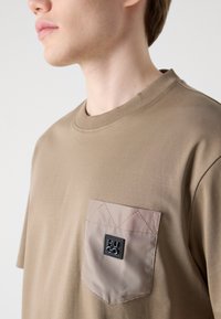 Tricou maro cu mânecă scurtă, având o textură subtilă, prezentând un buzunar patch de culoare bej cu un model în romb și un eticheta cu logo negru.