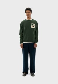 Felpa verde con una grafica di un albero textured sul petto, colletto e polsini a costine; abbinata a pantaloni blu navy larghi e scarpe scure.