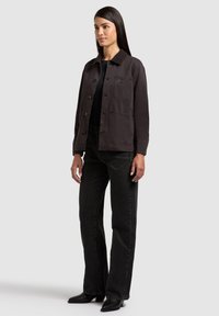 Femme debout portant une veste noire à boutons avec col, un haut noir, un jean taille haute noir et des bottes noires à bout pointu sur fond gris.