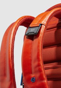 Rode leren rugzakriem met een gestructureerde stofvoering. Bevat een zwart label met "M.P." en een blauwe accentclip. Ritsdetail zichtbaar.