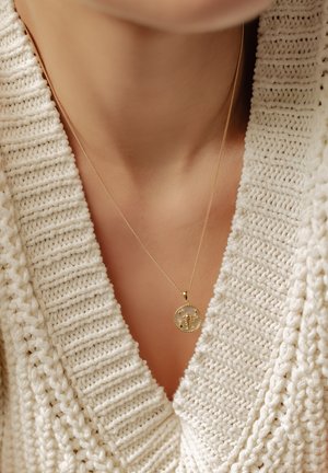 Gold pendant necklace on a thin chain, featuring a circular charm with floral detailing, worn over a textured, cream-colored sweater.