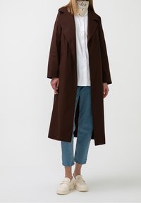 Person som bär en lång brun kappa, vit skjorta, ljusblå cropped jeans, krämfärgade klumpiga loafers och en beige mönstrad halsduk.