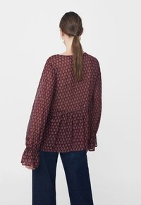 Blusa trasparente bordeaux con un motivo geometrico ripetuto, maniche lunghe con polsini arricciati e orlo arricciato. Abbinata a jeans blu scuri.