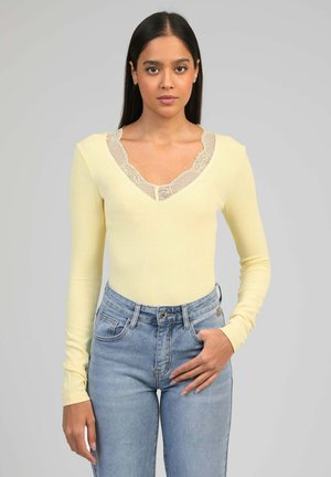 Jeune femme debout portant un haut à manches longues jaune pâle avec un col en dentelle et un jean taille haute bleu clair, sur un fond gris.