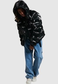 Geacă puffer neagră cu finisaj lucios, având imprimeuri albe cu logo. Combinată cu blugi evazați de culoare albastru deschis și adidași albi.