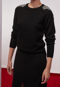 Pull noir avec un col arrondi, des poignets côtelés et des accents bijoux sur les épaules. Porté avec une jupe noire dotée d'une fente latérale.