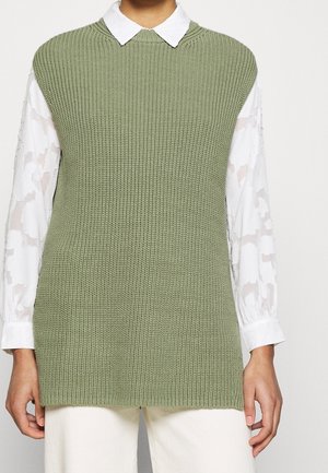 Person iført lysegrøn ærmeløs strikket sweatervest over hvid langærmet bluse med blomsterprint og hvide bukser.