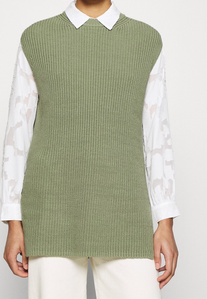 Personne portant un gilet sans manches en tricot vert clair sur un chemisier blanc à manches longues avec des motifs floraux et un pantalon blanc.