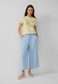  Lichtgele T-shirt met een gedrukt ontwerp, gecombineerd met losse lichtblauwe wijde broek en witte sandalen met bandjes. Glad stoftextuur.