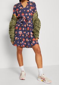 Blommönstrad klänning i marinblått med vita och rosa blommor, i lagerdesign, V-ringning och knytmidja, kombinerad med en olivgrön bomberjacka och sneakers.