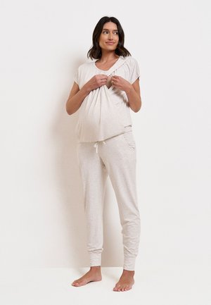 Zwangere vrouw in lichtgrijze loungewear gebruikt haar handen om het voedingsluik in het shirt te openen, staand op blote voeten tegen een effen achtergrond.