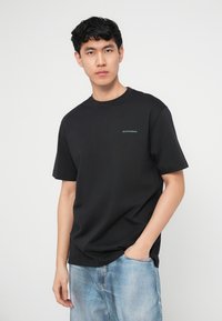 Νέος άνδρας που φοράει μαύρη κοντομάνικη μπλούζα με μικρό κείμενο στο στήθος και ανοιχτόχρωμα τζιν τζιν, με το ένα χέρι στην τσέπη, ουδέτερο φόντο.