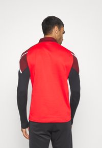 Rode sportieve shirt met lange zwarte mouwen, met een geometrisch patroon langs de schouders en een hoge kraag voor bedekking.