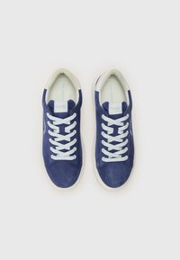 Sapatilhas de camurça azul com atacadores azul claro, solas de borracha brancas e um forro branco em contraste. Apresenta o logótipo da marca de lado.