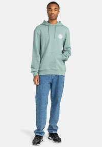 Mint green hooded sweatshirt with a front pocket, ribbed cuffs, and a circular logo on the chest, paired with blue denim jeans and black sneakers.