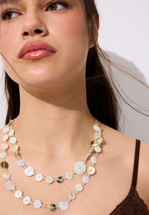 Detailná fotografia ženy nosiacej vrstvené biele a zlaté náhrdelníky s diskami a priehľadnými korálkami, tmavé vlasy zviazané dozadu a viditeľný hnedý čipkovaný popruh topu.