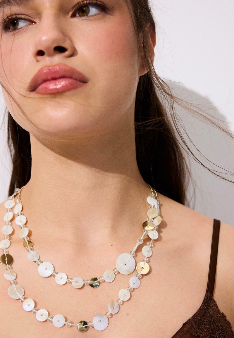 Gros plan d'une femme portant des colliers superposés blancs et dorés avec des perles transparentes, cheveux foncés attachés en arrière et une bretelle visible d'un haut en dentelle marron.