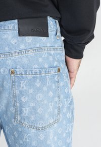 Los jeans de mezclilla azul claro presentan un sutil patrón de monograma en toda la prenda, una etiqueta de cuero negro en la cinturilla y detalles en tono dorado.