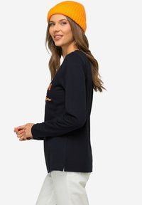 Granatowa koszula z długim rękawem i rozcięciami po bokach, z pomarańczowym napisem. Połączona z jaskrawym pomarańczowym czapką zdzierganą. Widoczne białe spodnie.