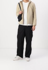 Osoba ubrana w beżową kurtkę z kapturem, białą koszulkę, czarne spodnie typu cargo, białe sneakersy i czarną torebkę na ramię na białym tle.