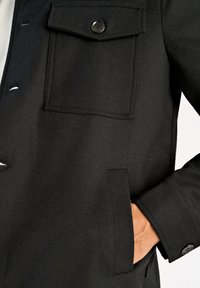 Svart jacka med texturerat tyg, som har en bröstficka, sidfickor och knäppning. Designen har en slank, skräddarsydd form.