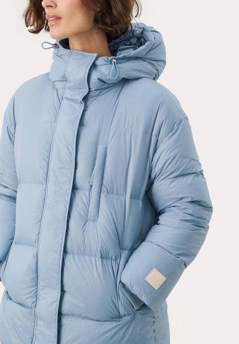 Helle blaue Pufferjacke mit hohem Kragen, Reißverschluss vorne und einer kleinen Brusttasche. Mit gerippten Bündchen und einem Logo-Patch am Ärmel.