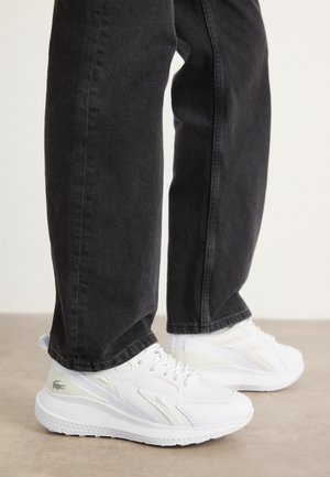 Zapatillas blancas con un diseño texturizado, que presentan un acento en verde claro, combinadas con jeans oscuros. Los zapatos tienen una suela ligera y cordones.