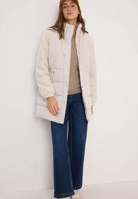 Veste rembourrée en crème et gris clair, avec une texture en polaire sur le dessus, des boutons-pression et des poignets élastiques. Associée à un jean large bleu.