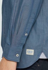 Chemise rayée bleue avec col boutonné, manches longues et étiquette sur l'ourlet. Texture lisse avec des accents de boutons.