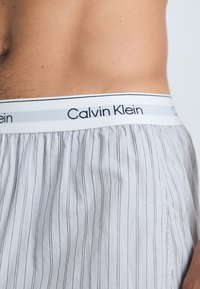 Vyrų torso, apsirengusio baltomis Calvin Klein bokseriu kelnaitėmis su plonomis vertikaliomis juodomis juostelėmis, matoma liemenių logotipas ir šoninės siūlės detalė.
