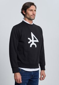 Hombre con sudadera negra con un gran símbolo abstracto blanco en el frente, camiseta blanca debajo y jeans azules, mirando hacia la derecha.