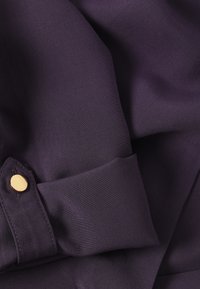 Tissu violet foncé avec un revers plié orné d'un seul bouton rond en or, montrant les détails de la texture et des coutures du matériau.