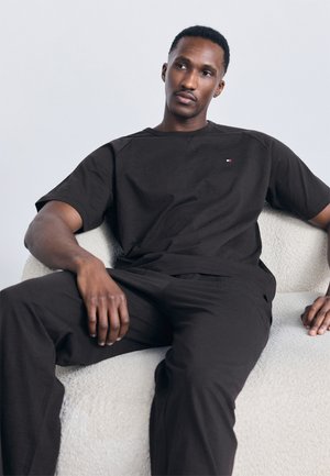 Bărbat purtând tricou negru și pantaloni, stând relaxat într-un fotoliu alb cu textură, pe un fundal deschis simplu.