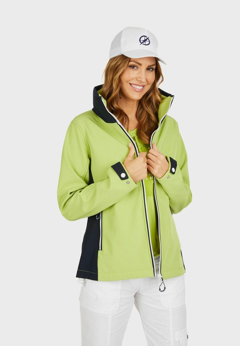 Groene softshell jack met een hoge kraag, marineblauwe accenten en zilveren ritsen. Draagt over een lichte top. Witte pet met een logo.
