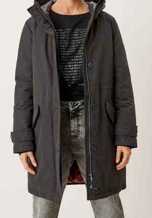 Osoba nosící dlouhý černý kabát s knoflíky a zipy, pod ním černé tričko s bílým textem a šedé džíny s vypraným efektem.