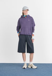 Sudadera con capucha morada y bolsillo frontal, gorra gris, mangas largas de patrón azul y naranja, pantalones cortos oscuros y zapatillas deportivas ligeras.