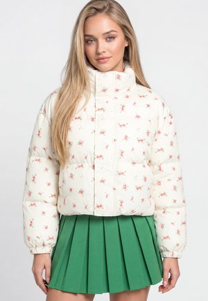 Vrouw met lang blond haar draagt een witte bloemen-gebloemde donsjas en een groene geplooide rok, staand tegen een egale lichte achtergrond.