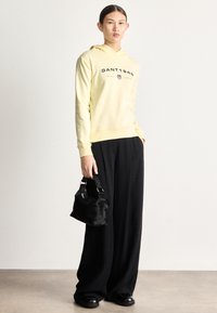 Žltá mikina s kapucňou a potlačeným logom, kombinovaná so širokými čiernymi nohavicami a malou čiernou kabelkou z umelého kožušiny. Hladká textúra, ležérny strih.